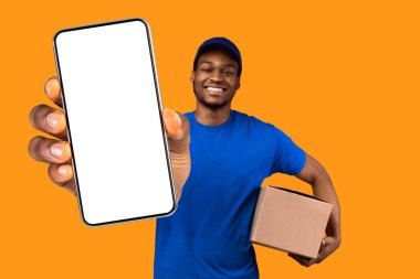 En İyi Teslimat Hizmeti Konsepti. Mavi şapkalı ve üniformalı gülümseyen siyah kurye elinde karton kutuyla sarı turuncu stüdyo arka planında izole bir şekilde duran baş parmak hareketlerini gösteriyor.