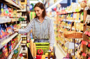 Satış, Alışveriş, Tüketim ve İnsanlar Konsepti. Gülümseyen genç bir kadın, elinde bir sepet dolusu ekolojik ürünle markette rafların yanında yürüyor, markette sağlıklı yiyecekleri seçiyor.
