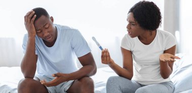 İstenmeyen hamilelik ve ilişkiler. Hamile olan kızgın siyah kadın şok olmuş erkek arkadaşına bağırıyor. Kafasına dokunuyor. Afro-Amerikan çift evde yatakta oturuyor.