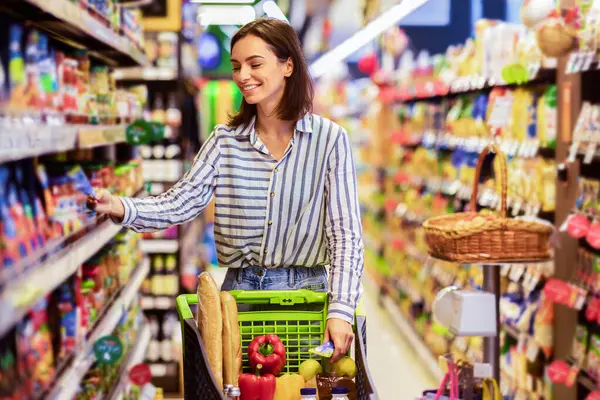 Her gün alışveriş konsepti. Gülümseyen Genç Ev Kadını Markette Koridorlar Arasında Trolley Arabası İtiyor. Pozitif kadın yemek alıyor, raftan ürün alıyor. Tüketim ve Yaşam Tarzı