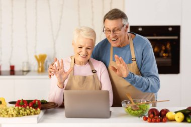 Joyful Senior Couple Video Calling Using Laptop Waving Hands To Computer, Communicating With Distant Friends Online Standing In Kitchen At Home. Modern Remote Communication, Video Call Concept