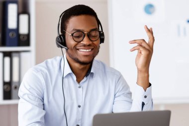 Pozitif Afro-Amerikalı genç adam ofiste online iş konferansı yapıyor, dizüstü bilgisayar ve kulaklık kullanıyor, müşteriyle veya iş ortağıyla neşeli bir şekilde konuşuyor, el kol hareketi yapıyor ve gülümsüyor, kopyalama alanı kopyalıyor.