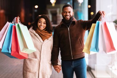 Alışveriş zamanı geldi. Neşeli zenci erkek ve kadın portresi. Renkli çantalar gösteriyorlar. Geceleri dışarıda dolaşıyorlar. Afro-Amerikalı bir çift dükkan vitrininin yanında bekliyor.