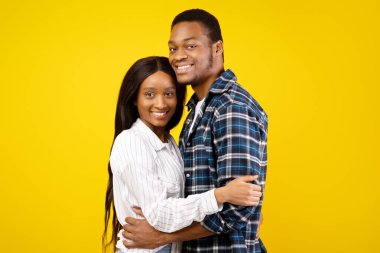 Aşık çiftlerin romantik ilişkisi ve iki kişilik Covid-19 karantinası. Gülümseyen Afro-Amerikan erkek ve bayan, sıradan giyinmiş, sarılıyor, sarı arka planda izole edilmiş, stüdyo çekimi, kopyalama alanı