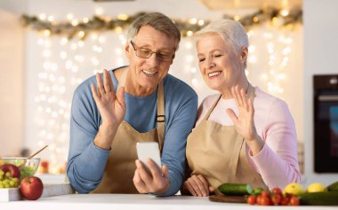 Modern Mutfak 'ta Noel' i kutlamak için akıllı telefondan el sallayan mutlu yaşlı çift. Uzaktan Aile İletişimi. Üst Düzey Eşler Uzaktan Akrabaları Telefon Uygulaması Kullanarak Arıyor