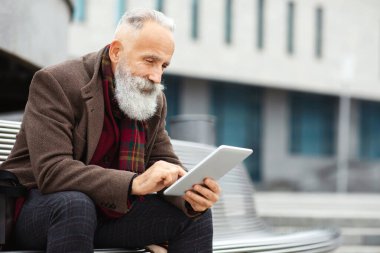 Tarz sahibi gri saçlı yaşlı bir adam ofis binasının yanındaki bankta oturuyor, dijital tablet kullanıyor, ajandasını kontrol ediyor, kopyalama alanını kopyalıyor. Yaşlı adam sokakta ped kullanıyor, dinlenirken web sörfü yapıyor.