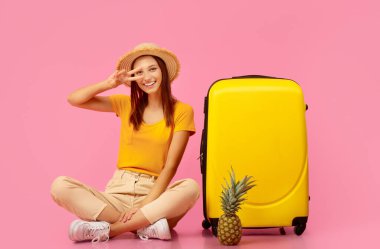Ananaslı mutlu bir kadın bavulun yanında oturmuş pembe stüdyo arka planında poz veriyor. Kaygısız genç bayan egzotik sıcak ülkelere seyahat ediyor, ateşli anlaşmalar.
