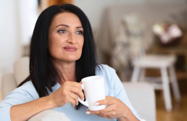 Weekend Leisure. Portrait of smiling mature woman drinking hot coffee, holding cup and sitting on the couch in living room. Happy lady enjoying good morning. Resting And Relax Concept