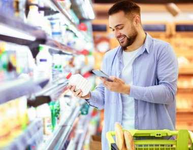 Gülümseyen genç adam marketteki raflardan süt ürünleri alıyor, elinde şişe ve akıllı telefon, müşteriler barkodu tarıyor cep telefonuyla, el arabası ile yürüyor.