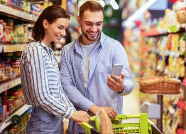 Satış ve indirim arıyorum. Süpermarkette Neşeli Milenyum Ailesinin Portresi. Birlikte alışveriş yapan genç bir çift markette arabayla gezen bir telefon uygulaması kullanıyor.