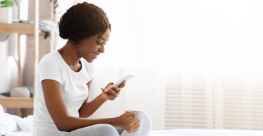 Kahveli neşeli siyah kadın yatakta oturuyor ve cep telefonu, yan görüş, fotokopi odası kullanıyor. Gülümseyen Afrikalı Amerikalı kadın. Sabahları akıllı telefondan sohbet ediyor. Yatak odasının içinde.