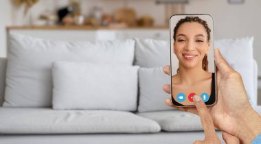 Bir kişi video bağlantısı kurmak için akıllı telefon kullanıyor. Sıradan bir ev ortamı sergiliyor. Ekrandaki kişi mutlu ve sohbete odaklanmış görünüyor..