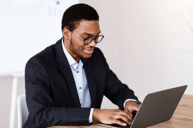 Resmi takım elbiseli gülümseyen siyah yönetici dizüstü bilgisayarla çalışıyor, gözlük takıyor, ofis içi, fotokopi odası. Başarılı bin yıllık Afro-Amerikan iş adamı iş projesi üzerinde çalışıyor.