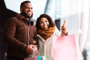 Buraya bak. Heyecanlı, Afro-Amerikalı güzel bir bayan erkeğine moda kıyafetlerini gösteriyor, mağazanın camını işaret ediyor, akşamları alışveriş merkezinin yakınında yürüyor. Satış, İndirim, Perakende, Satın Alım