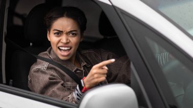 Angry, young, pissed off, displeased, aggressive african american woman shouting on drivers in front of her and points finger, driving car. Road traffic jam, negative human emotions and scandal