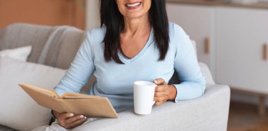 Leisure, Rest And Relax Concept. Portrait of smiling mature woman reading book and holding mug of coffee, sitting on the sofa in living room in her apartment, enjoying weekend and looking at camera