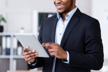 Teknolojiler iş başında. Siyah takım elbiseli mutlu Afro-Amerikan genç işadamı ofiste dijital tablet kullanıyor, internette çalışıyor, pazarlama raporlarını okuyor, sunuma hazırlanıyor, uzayı kopyalıyor.