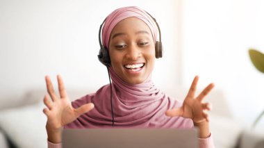 Başörtüsü takmış, kulaklık takan çekici Müslüman bir kadın evde dizüstü bilgisayarla iletişim kuruyor. Geleneksel türbanlı sevimli bayan öğretmen web kamerası üzerine uzaktan ders veriyor.