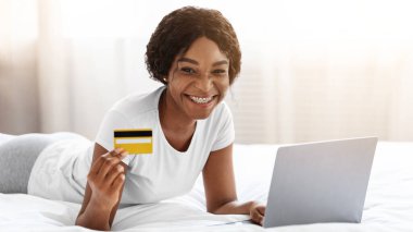 Evden internet alışverişi, e-ticaret konsepti. Neşeli genç Afro-Amerikalı kadın evde yatıp internetten kıyafet ya da ürün alıyor, kredi kartıyla ödüyor, dizüstü bilgisayarla, boş alanla.