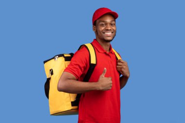 En İyi Teslimat Hizmeti Konsepti. Kırmızı üniformalı ve termal çantalı özgüvenli gülümseyen siyah kurye portresi baş parmak işareti yapıyor ve mavi arka planda izole edilmiş kameraya bakıyor.