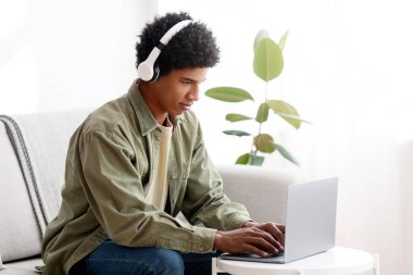 Black teen guy watching educational webinar on laptop, wearing headphones, taking part in web lesson at home. Young teenager having online meeting with university tutor