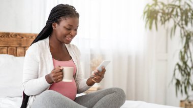 Hamilelik sırasında boş zaman. Siyah bir bayanın yatakta akıllı telefonuyla dinlenmesini, çay içmesini, internete göz atmasını ve sıcak içeceklerin tadını çıkarmasını, hamile Afrikalı bir kadının evde dinlenmesini, boşluğu kopyalamasını bekliyorum.