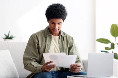 Happy black guy learning online from home, writing coursework, holding documents near laptop. African American teen student consulting with his teacher about educational project