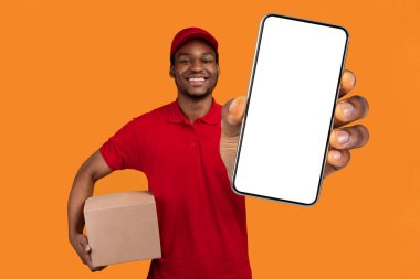 Kolay ödeme. Kırmızı şapkalı ve üniformalı gülümseyen Afro-Amerikan erkek kurye elinde kredi kartı ve karton kutu, paket taşıyor, turuncu stüdyo arka planında tek başına duruyor.