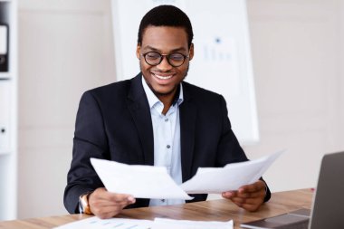 Gözlüklü neşeli siyah yönetici pazarlama raporlarını okuyor ve gülümsüyor, işyerinde laptopun önünde oturuyor, Afro-Amerikan işadamı evrak işleriyle uğraşıyor, kopyalama alanı yaratıyor.