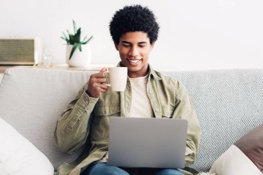E-öğrenme konsepti. Yakışıklı siyah erkek öğrenci dizüstü bilgisayar kullanıyor ve evde sıcak kahve içiyor, panorama. Çevrimiçi konferans dinleyen mutlu genç adam, web kamerasından öğretmenle iletişim kuruyor.
