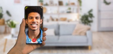 Çevrimiçi bir iletişim oturumu sırasında gülümseyen bir kişiyi gösteren bir akıllı telefon tutan bir kişi. Konforlu evde şık bir kanepe ve dekoratif bitkiler var..