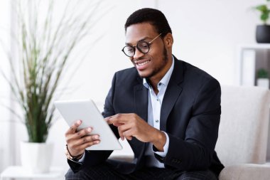 İş dünyasındaki modern teknolojiler. Mutlu Afro-Amerikan takım elbiseli genç işadamı ofiste dijital tablet kullanıyor, internette çalışıyor, pazarlama raporlarını okuyor, sunuma hazırlanıyor, uzay kopyaları kopyalıyor.