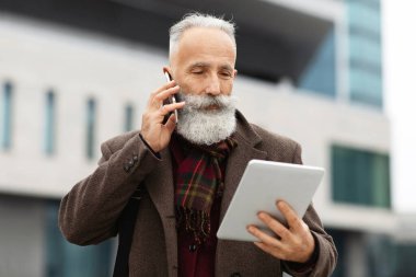 Ciddi, gri saçlı, uzun sakallı bir adam dijital tablet ekranına bakıyor ve cep telefonuyla konuşuyor. Sokakta yürürken iş görüşmesi yapıyor.