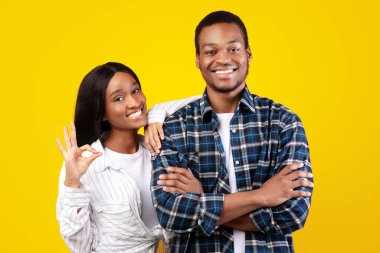 İyi bir müşteri değerlendirmesi, müşteri tavsiyesi. Genç, neşeli Afro-Amerikan kadın elini erkeğin omzuna koydu ve el işaretini gösterdi, sarı arka planda izole edilmiş, stüdyo çekimi.
