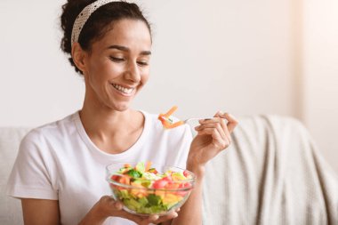 Elinde sağlıklı salata ve çatalla gülümseyen, evdeki kanepede oturup fotokopi çeken güzel bir genç kadın. Sağlıklı genç esmer kadın öğle yemeğinde sebze yiyor. Diyet kavramı
