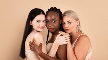 Farklı vücut ve ten tiplerine sahip üç farklı ırktan kadın grubu bej rengi stüdyo arka planında birlikte poz veriyorlar. İç çamaşırlı güzel hanımlar kameraya sarılıyor ve ona bakıyor.