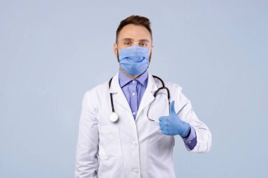 Laboratuvar önlüklü, koruyucu maskeli ve lastik eldivenli bir doktor gri stüdyo geçmişinde baş parmak hareketlerini gösteriyor. Genel pratisyen doktor covid-19 salgını sırasında koruma takıyor.
