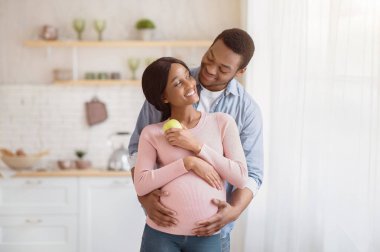 Pozitif hamile siyah çift, mutfakta pencerenin önünde yan yana dururken birbirlerine sarılıyorlar. Elmalı hamile bir kadın ve sevgi dolu kocası evde birbirlerine sevgi gösteriyorlar.