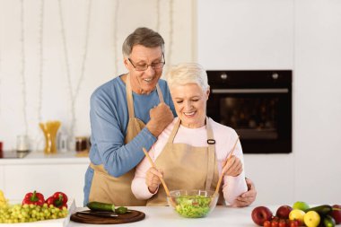 Gülümseyen son sınıf çifti, modern mutfakta birlikte yemek pişirmek için salata yapıyor. Yaşlı eşler öğle yemeği için sağlıklı sebze yemeği hazırlıyorlar. Diyet ve Sağlıklı Beslenme Yaşlılıkta