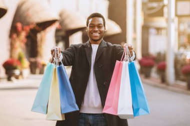 Satış sezonu başlıyor ve tatilden önce alışverişe çıkmak için Kara Cuma. Mutlu Milenyum Afrikalı Amerikalı adam alışveriş sonrası alışveriş merkezinin yakınındaki renkli çantaları gösteriyor.