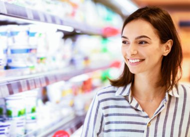 Perakende konsepti. Marketteki rafların yanında duran gülümseyen genç kadının yakın plan portresi. Neşeli Kadın Müşteri Süper Markette Temelleri Satıyor, Süt Ürünleri ile Koridora Bakıyor