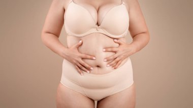 Obezite ve kilolu. Bej rengi stüdyo arka planında duran, iç çamaşırlı, iç çamaşırlı, yüz kızartıcı bir kadın, vücut utancından muzdarip.