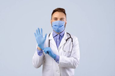Kendine güvenen maskeli genç erkek doktor gri stüdyo geçmişine eldiven takıyor. Koronavirüs salgını sırasında önlük giyen başarılı bir tıp çalışanı.