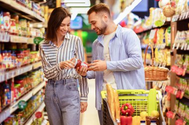 Sağlıklı beslenme. Genç, gülümseyen bir çift marketten alışveriş yapıyor. İki alıcının elinde cam kavanoz sos var ve etiketleri kontrol ediyor, yiyecek ürünlerini seçiyor, alışveriş arabasıyla ayakta duruyor.