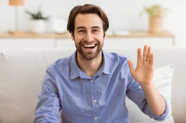 Hoş geldiniz. Mutlu adam el sallıyor ve kameraya gülümsüyor. Evdeki koltukta oturuyor. Neşeli adam el kol hareketi yapıyor, selam veriyor, içeride video görüşmesi yapıyor. Modern Uzaktan İletişim Konsepti