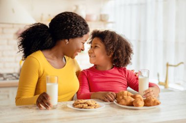 Mutfakta abur cubur yerken mutlu siyah anne ve kızı, neşeli Afrikalı Amerikalı annesi ve küçük çocuğu masada oturmuş kurabiye yiyor ve süt içiyor, bedava uzay boşluğu.