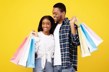 Boş zamanlarında indirimler, satışlar ve modern alışveriş. Şirin Afro-Amerikan erkek ve kadın sarılıp, renkli alışveriş çantaları taşıyorlar, sarı arka planda izole edilmiş, stüdyo çekimi, fotokopi alanı.