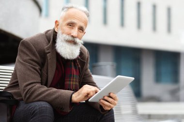 Modern yaşlı bir adam iş merkezinde bankta oturuyor ve dijital tablet kullanıyor, web sörfü veya sosyal medyayı kontrol ediyor, kopyalama alanı kullanıyor. Uzun gri sakallı yaşlı iş adamı.