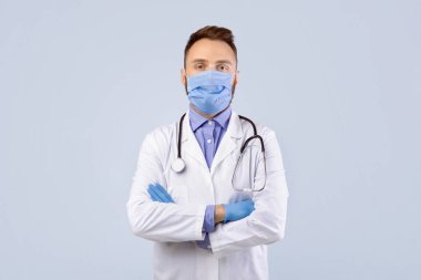 Profesyonel üniformalı erkek doktor, yüz maskesi ve lastik eldivenler gri stüdyo arka planında poz veriyor. Covid-19 salgını sırasında koruyucu takan kendinden emin bir tıp uygulayıcısı.
