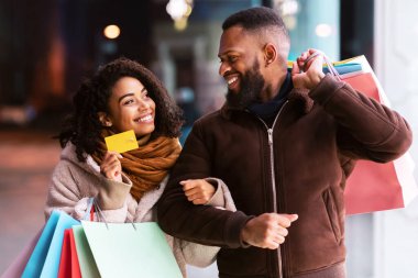 Bir şeyler alalım. Gülümseyen siyah kadının portresi bankamatik kartını bir erkeğe gösteriyor, alışveriş çantalarını tutuyor, akşamları alışveriş merkezinin yakınlarında yürüyor, geçmişi bulanık.